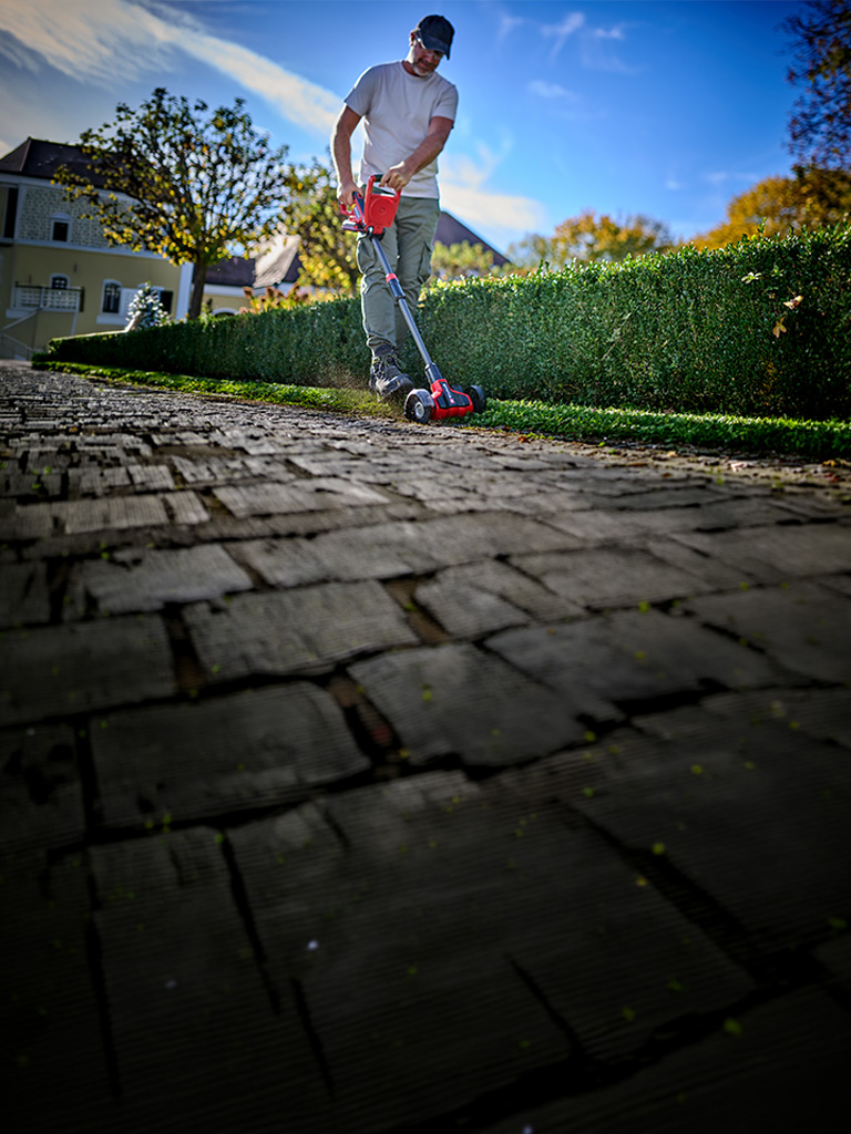 A man cleans paving joints along a hedge in the garden using the Einhell cordless patio cleaner.