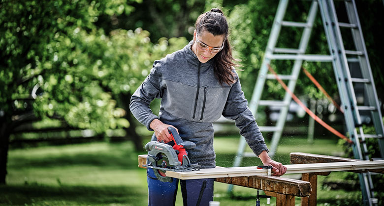 Woman wearing safety glasses cutting a wooden board outdoors with a red circular saw on two sawhorses.