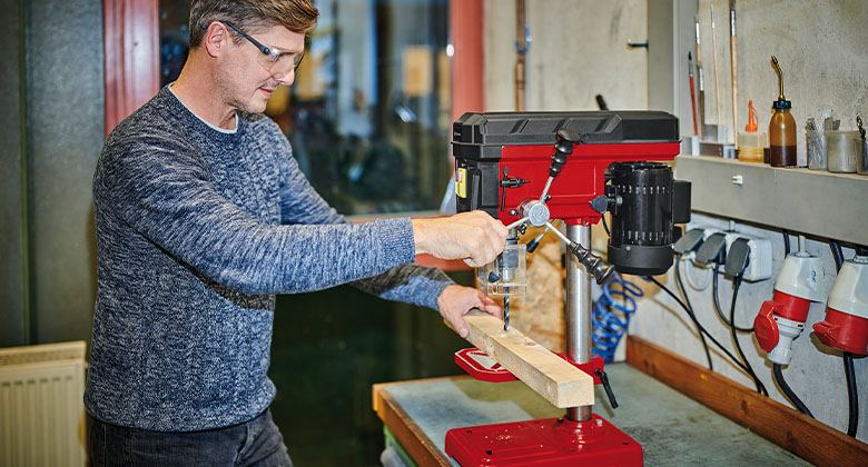 A man drilling into a wooden slat with a bench drill.