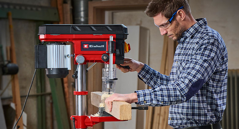 A man wearing safety goggles drilling a hole in a wooden slat with the help of a pillar drill.