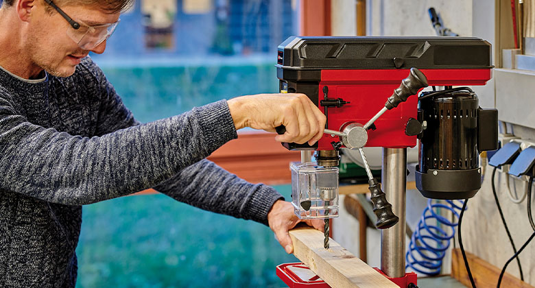 A man guiding the drill spindle of a bench drill downwards to drill into a wooden slat.