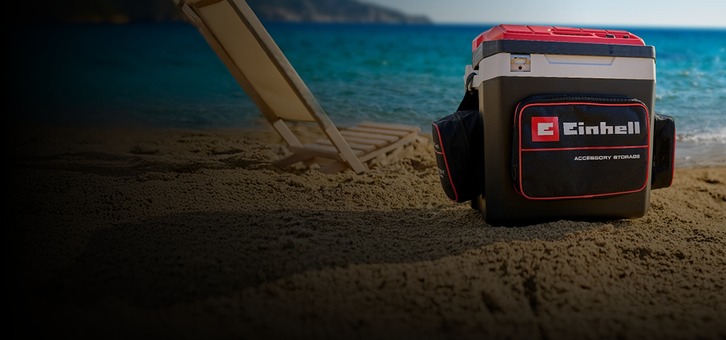 Das Bild zeigt eine Einhell-Kühlbox am Strand neben einer Liege. Im Hintergrund sieht man das blaue Meer und eine Küstenlandschaft.