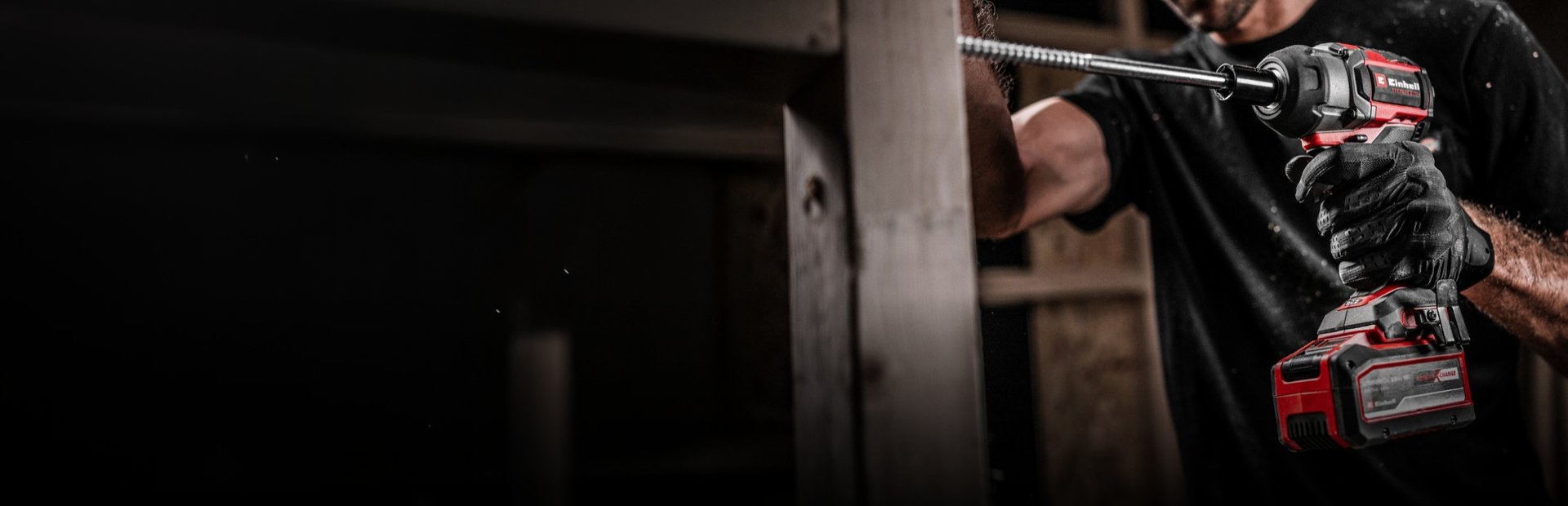 The image shows a man using a cordless Einhell impact wrench to drive a long screw into wood. He’s wearing gloves and the setting appears to be a construction site. 