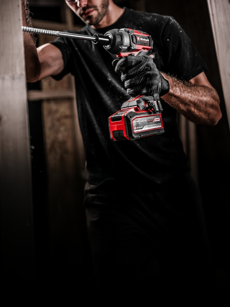 The image shows a man using a cordless Einhell impact wrench to drive a long screw into wood. He’s wearing gloves and the setting appears to be a construction site.