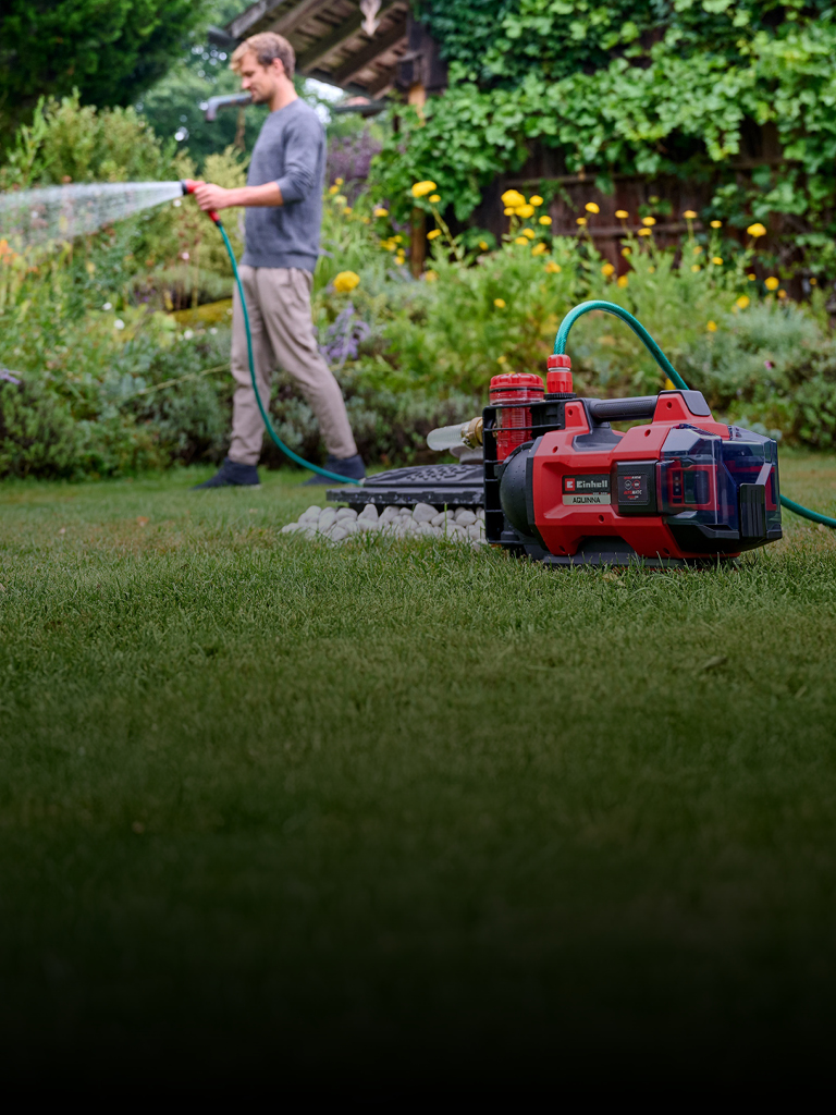 Ein Mann in Freizeitkleidung bewässert seinen Garten mit einem Schlauch, der mit einer roten und schwarzen Einhell-Akku-Wasserpumpe verbunden ist, die im Vordergrund auf dem Rasen steht. Im Hintergrund sind üppige grüne Pflanzen und gelbe Blumen zu sehen.