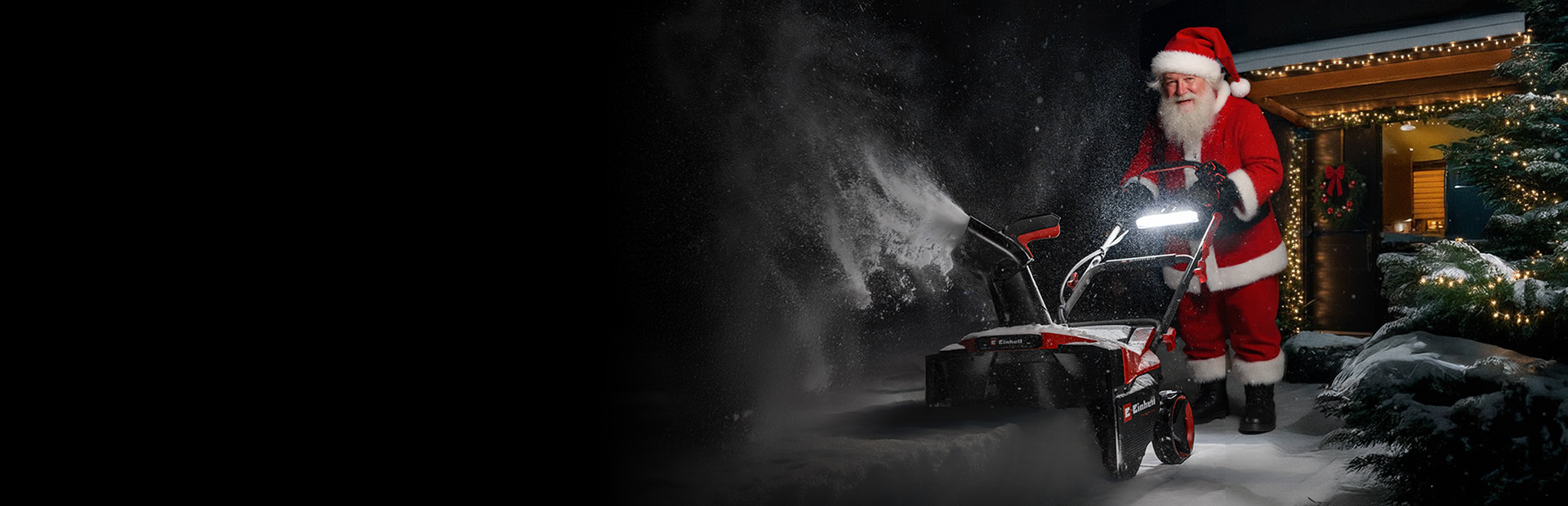 Eine als Weihnachtsmann verkleidete Person steht im Schnee vor einem mit Weihnachtsbeleuchtung geschmückten Haus und benutzt eine Schneefräse.