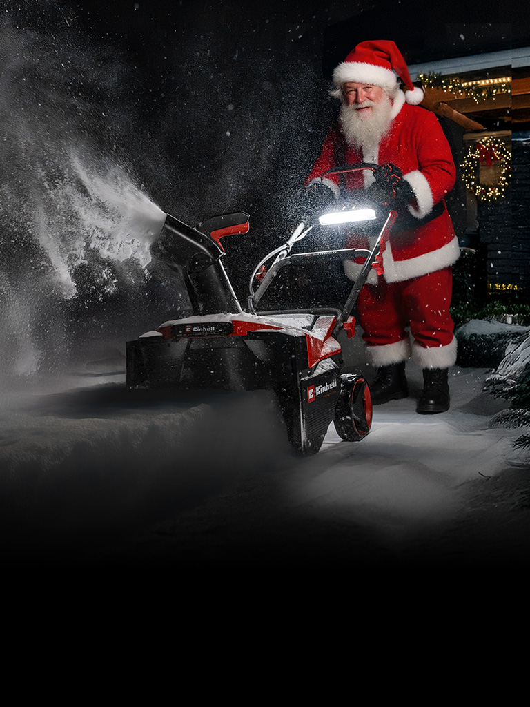 Eine als Weihnachtsmann verkleidete Person steht im Schnee vor einem mit Weihnachtsbeleuchtung geschmückten Haus und benutzt eine Schneefräse.