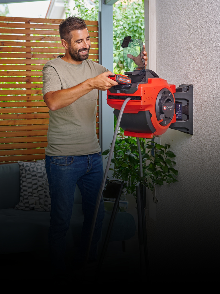 Un uomo sorridente inserisce una batteria in un avvolgitubo automatico rosso Einhell montato su una parete esterna. La scena è ambientata in un patio con una schermatura in legno e vegetazione sullo sfondo.