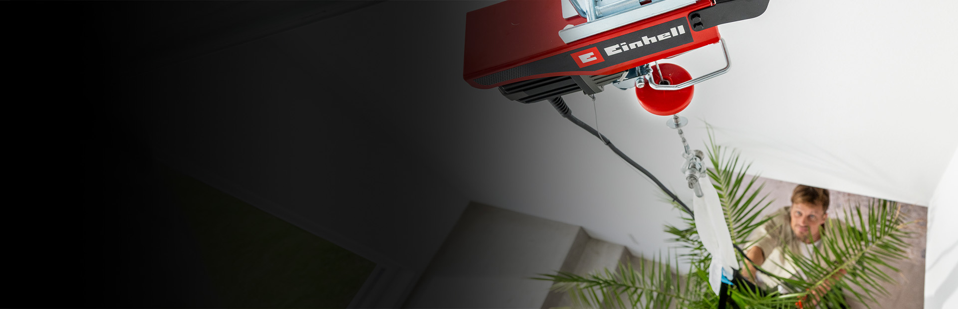 A person is holding a red cordless electric hoist while standing next to a large hanging plant in a modern indoor setting. The focus is on the hands and the device, with green leaves and support straps visible in the background.