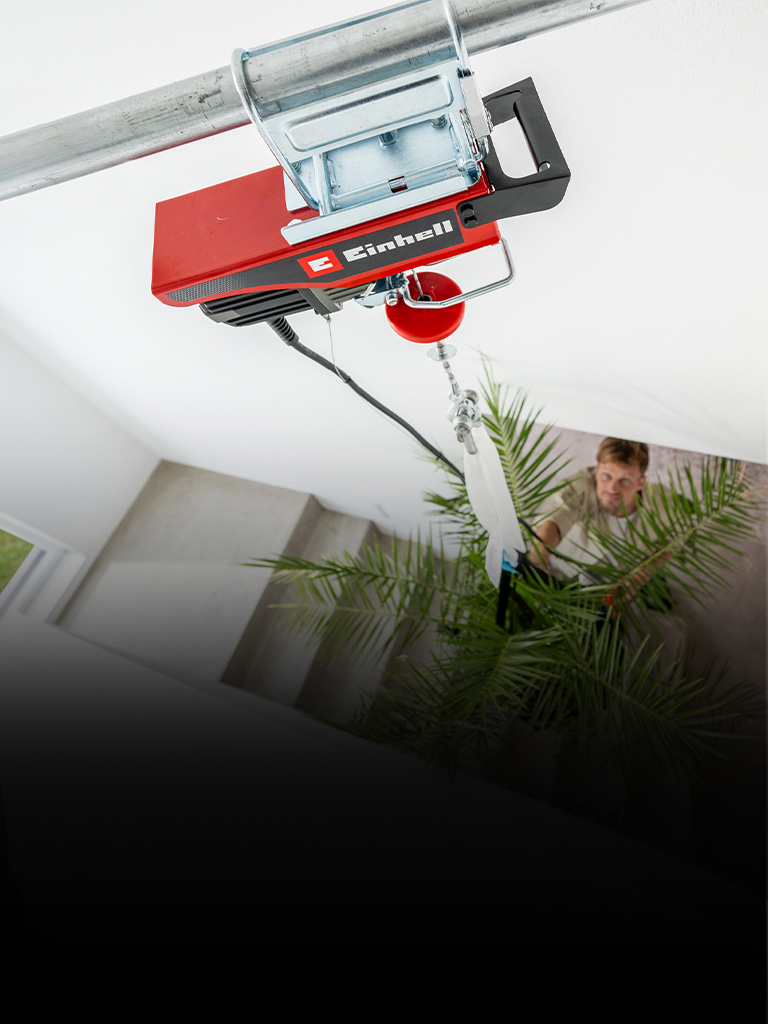 A person is holding a red cordless electric hoist while standing next to a large hanging plant in a modern indoor setting. The focus is on the hands and the device, with green leaves and support straps visible in the background.