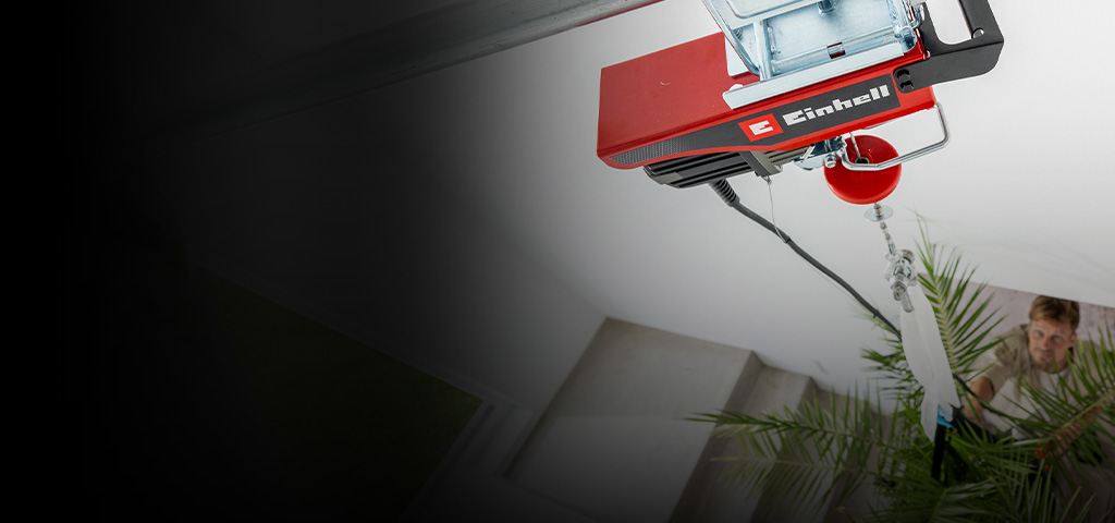 A person is holding a red cordless electric hoist while standing next to a large hanging plant in a modern indoor setting. The focus is on the hands and the device, with green leaves and support straps visible in the background.