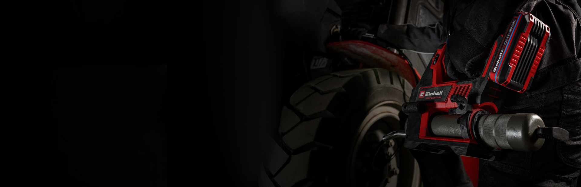 Eine Person führt Wartungsarbeiten an einem Fahrzeug durch und verwendet dabei eine rote, kabellose Fettpresse, während sie dunkle Arbeitskleidung trägt.