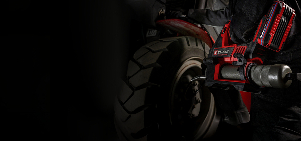 Eine Person führt Wartungsarbeiten an einem Fahrzeug durch und verwendet dabei eine rote, kabellose Fettpresse, während sie dunkle Arbeitskleidung trägt.