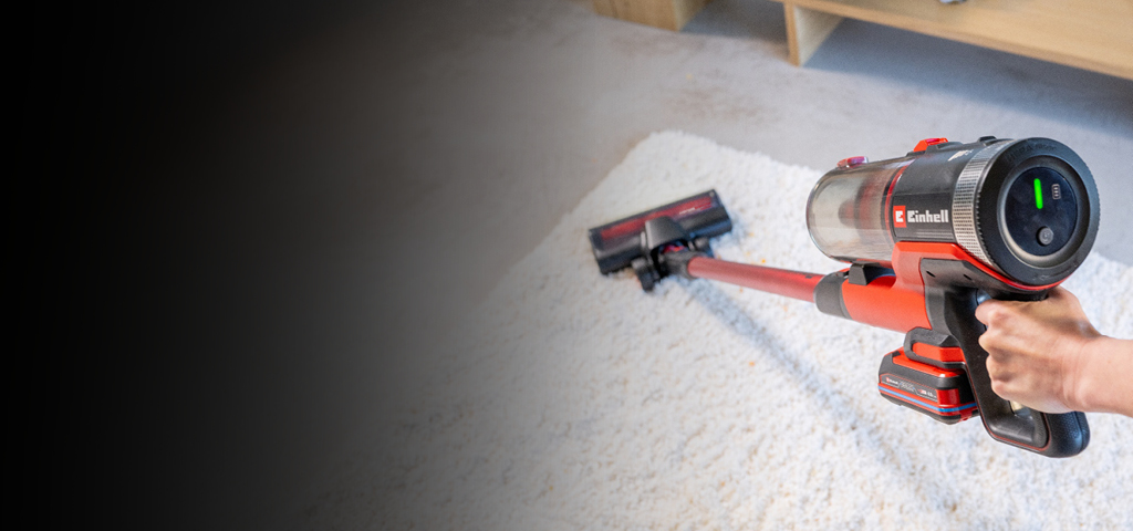 Das Bild zeigt einen kabellosen Staubsauger, der über einen hellen Teppich geführt wird. Der Staubsauger besitzt ein Display und auffällige rote Elemente, während im Hintergrund ein Teil eines Wohnzimmers mit Holzmöbeln erkennbar ist.