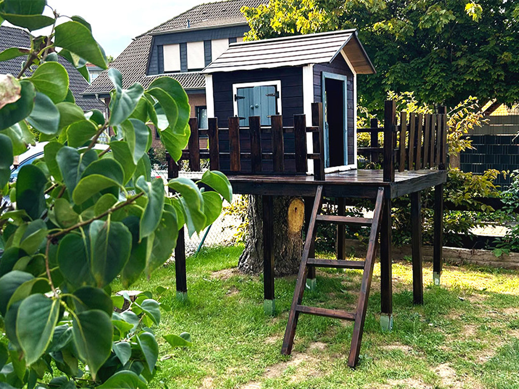 Homemade treehouse made of dark wood with a terrace and a small ladder, built on stilts around a tree in a garden.
