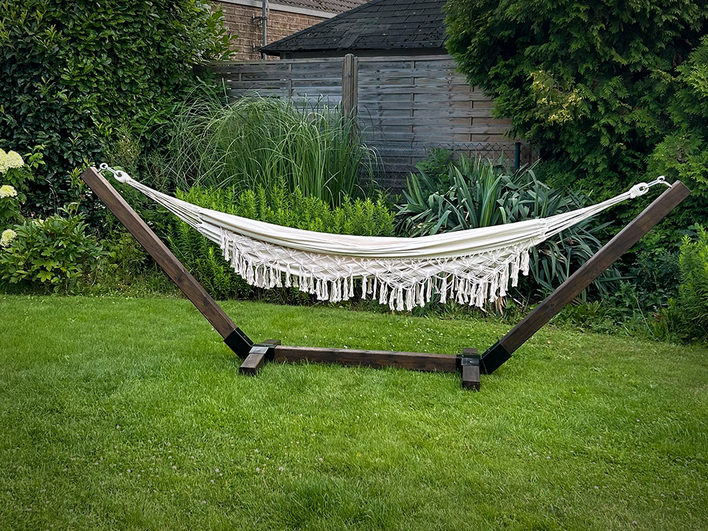 A cream-colored hammock with fringes hangs in a dark wooden frame on a lawn in a garden.