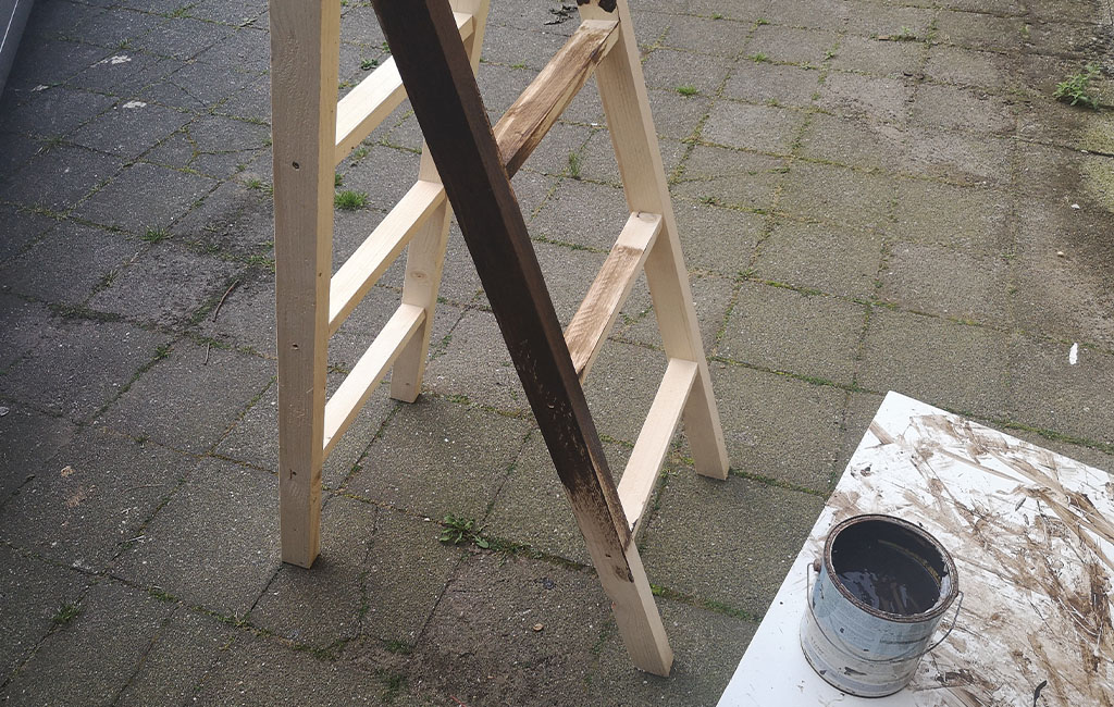 Eine Holzleiter wird teilweise mit dunkler Lasur gestrichen, eine offene Farbdose steht daneben.