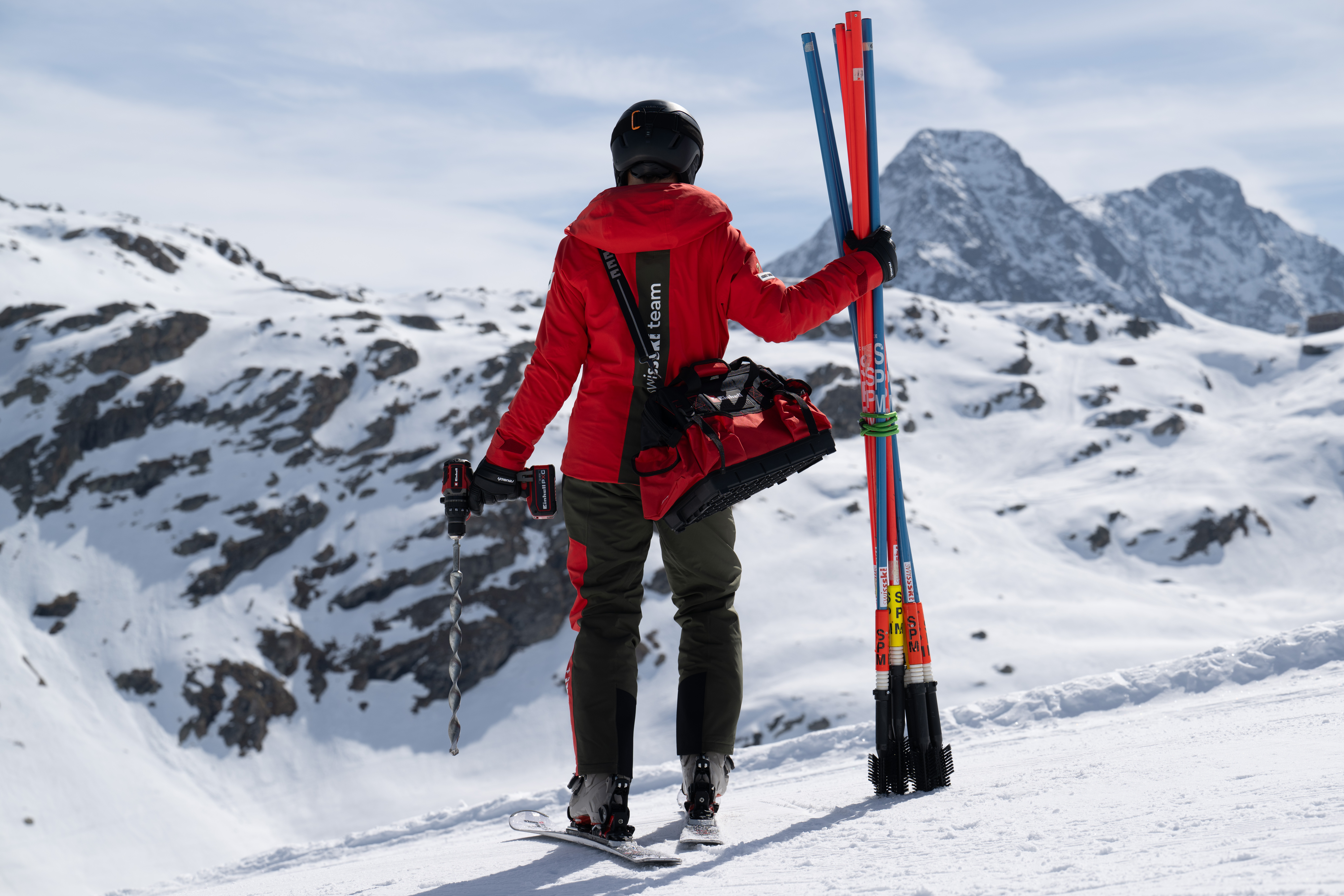 Ein Swiss-Ski-Team-Mitglied steht mit einem Einhell Akkubohrschrauber in der linken Hand und mehreren Slalomstangen in der rechten auf einem verschneiten Hang, mit Werkzeugtasche über der Schulter.